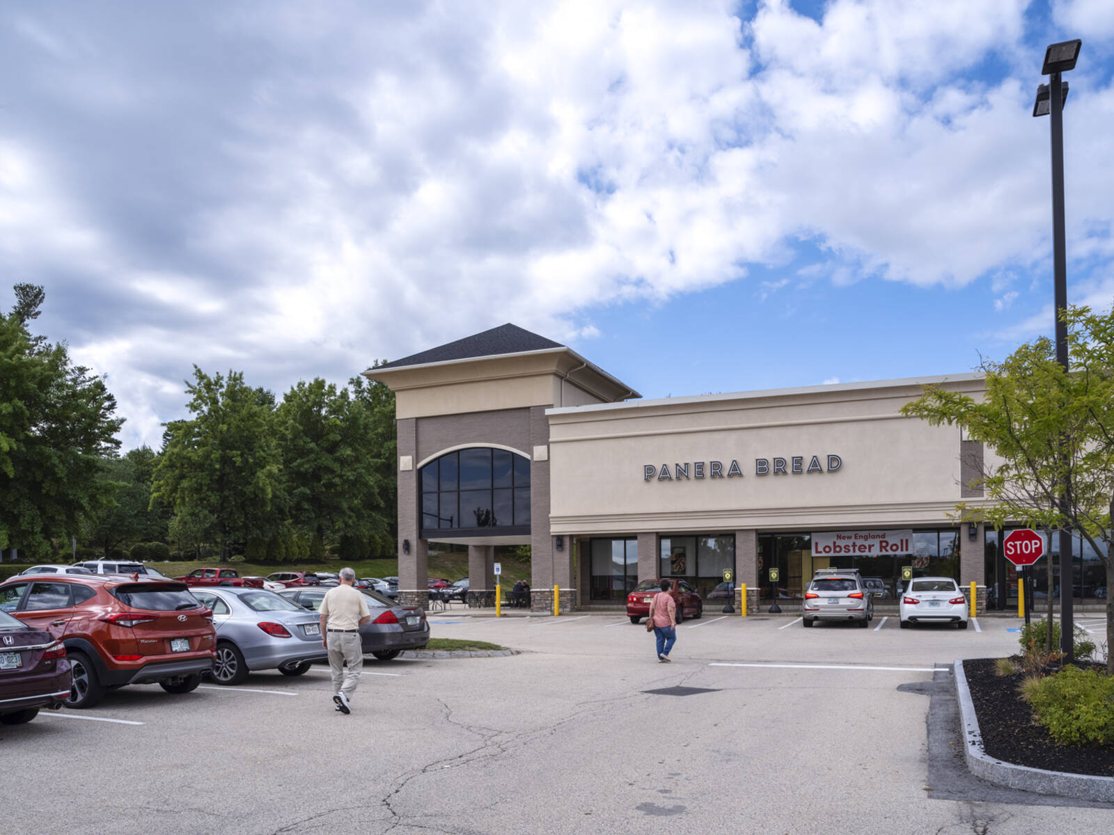 Pedestrians walk towards Panera Bread in a parking lot.