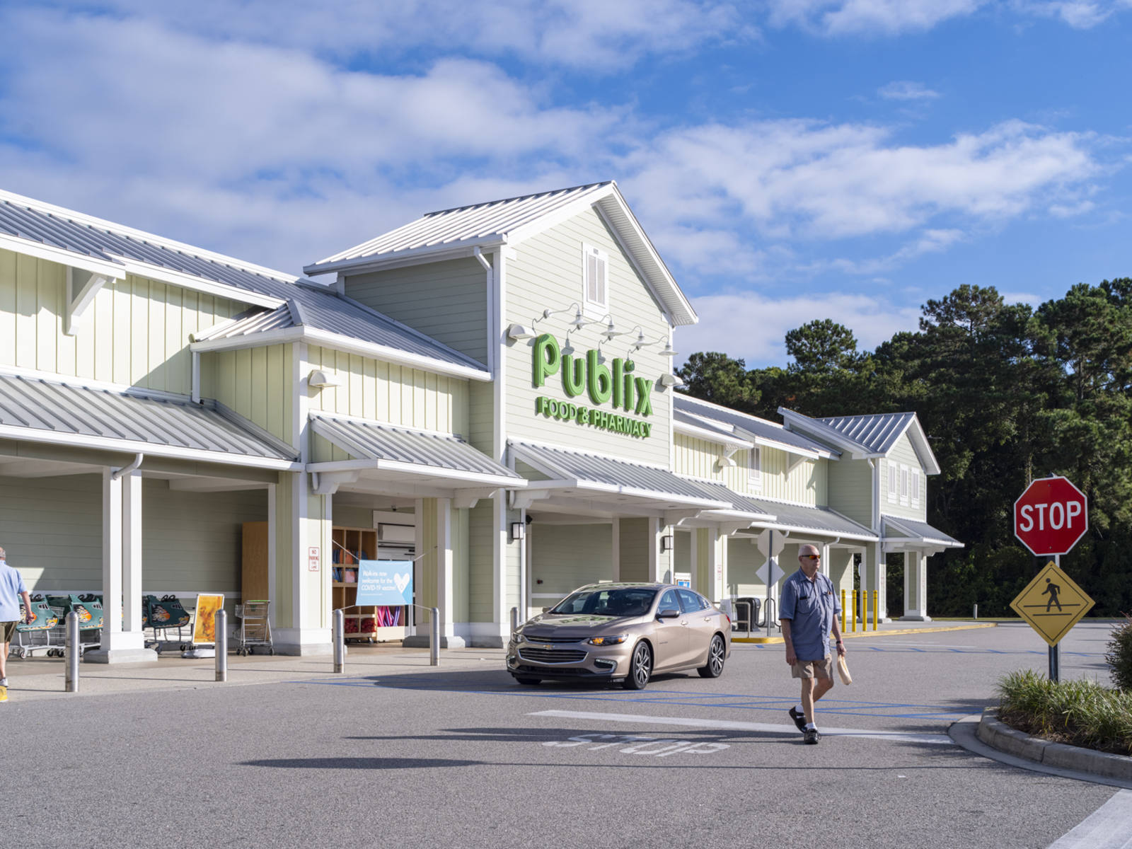 Person in blue shirt walking past tan car in front of entrance to Publix at outdoor shopping center