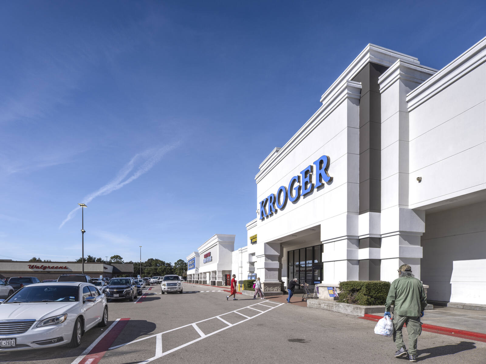 Pedestrians entering Kroger store with bags and carriages through busy parking lot.