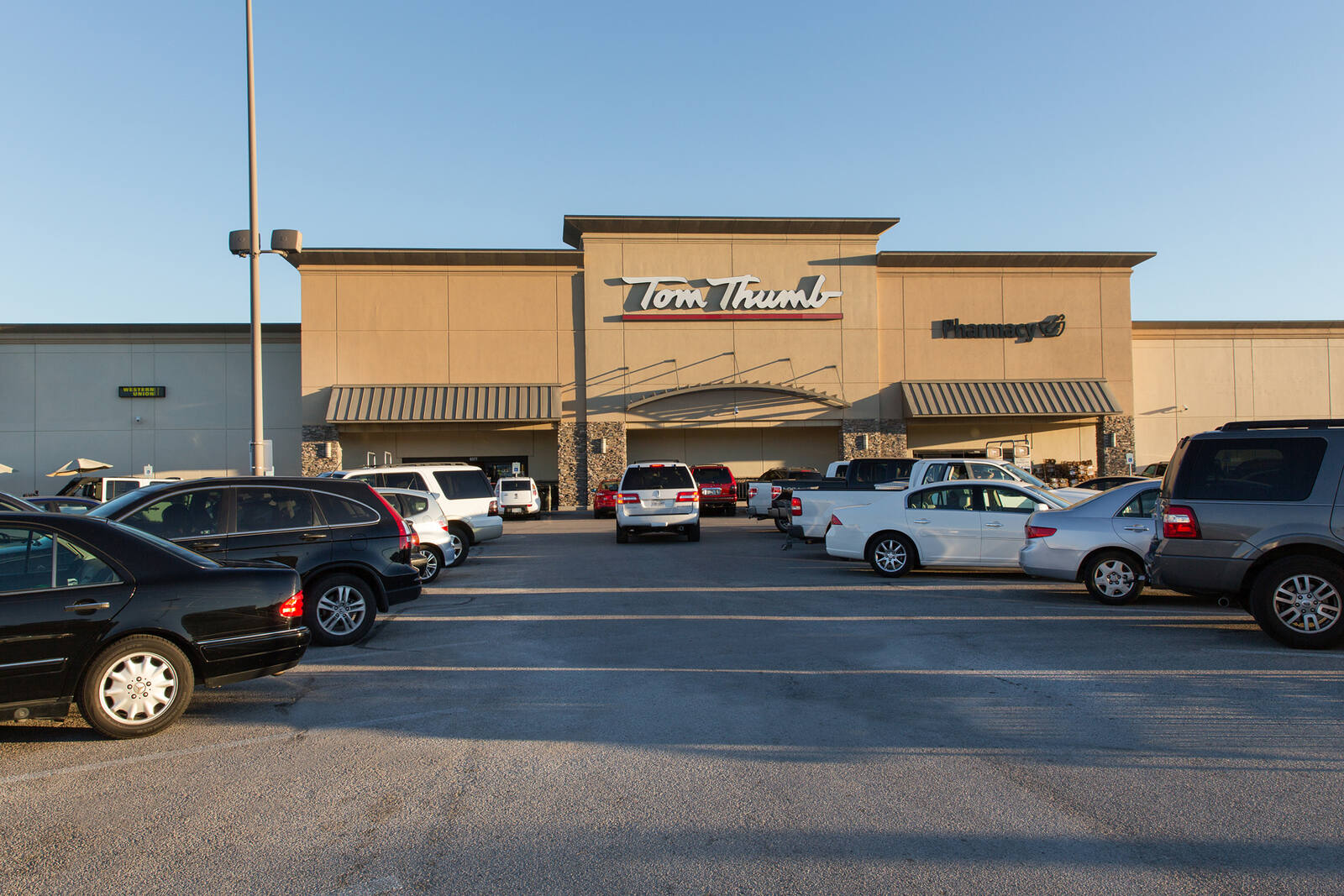 White cars driving through parking lot with cars parked on either side in front of Tom Thumb at Ridglea Plaza Shopping Center
